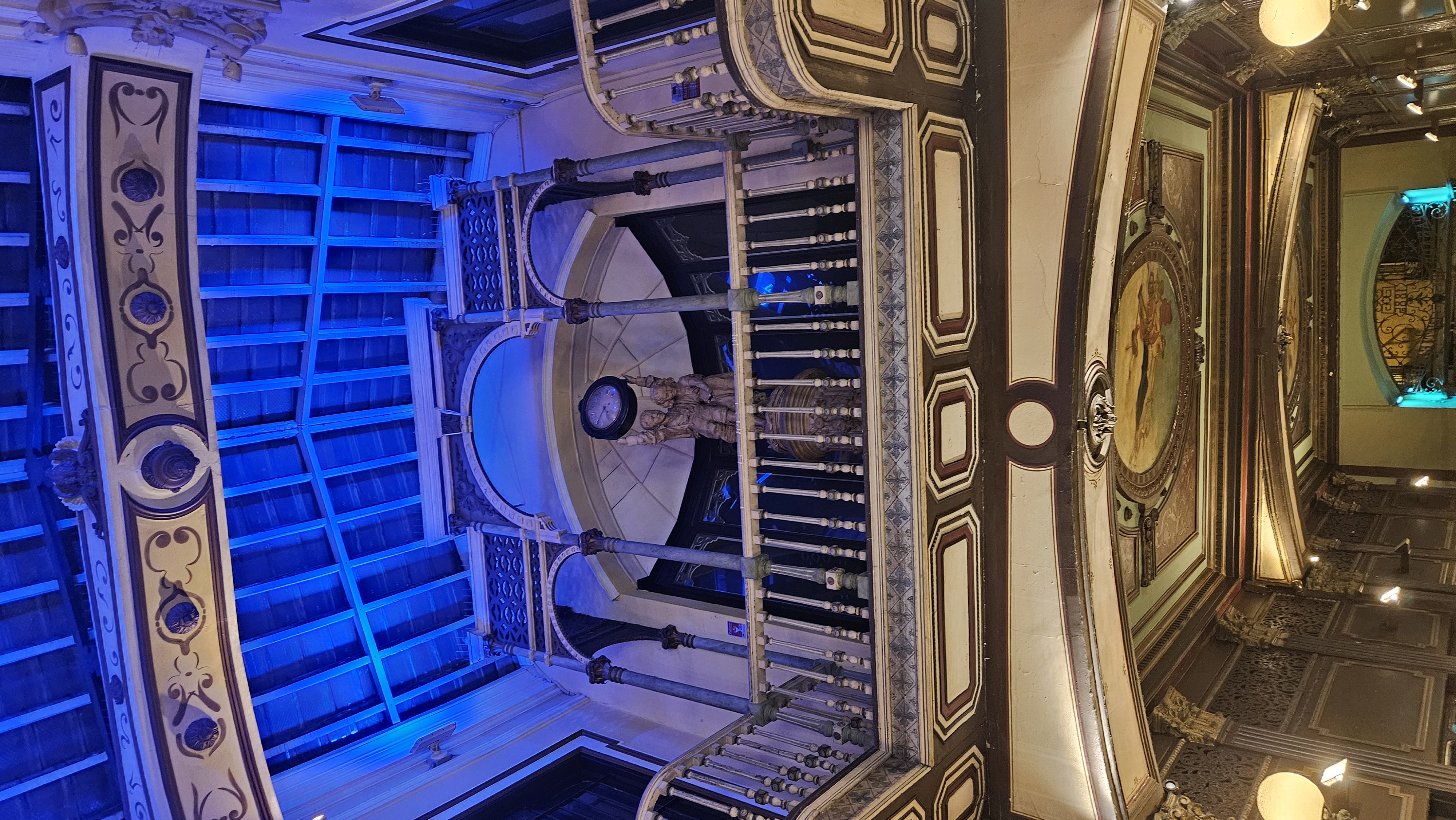 Fotografía del interior de un edificio histórico con una arquitectura ornamentada. En el centro destaca una escultura de dos figuras sosteniendo un reloj, ubicada en un balcón con columnas y barandillas decorativas. El techo de madera, iluminado con luces azules, y los techos abovedados con molduras y pinturas clásicas aportan un ambiente elegante y teatral al conjunto.