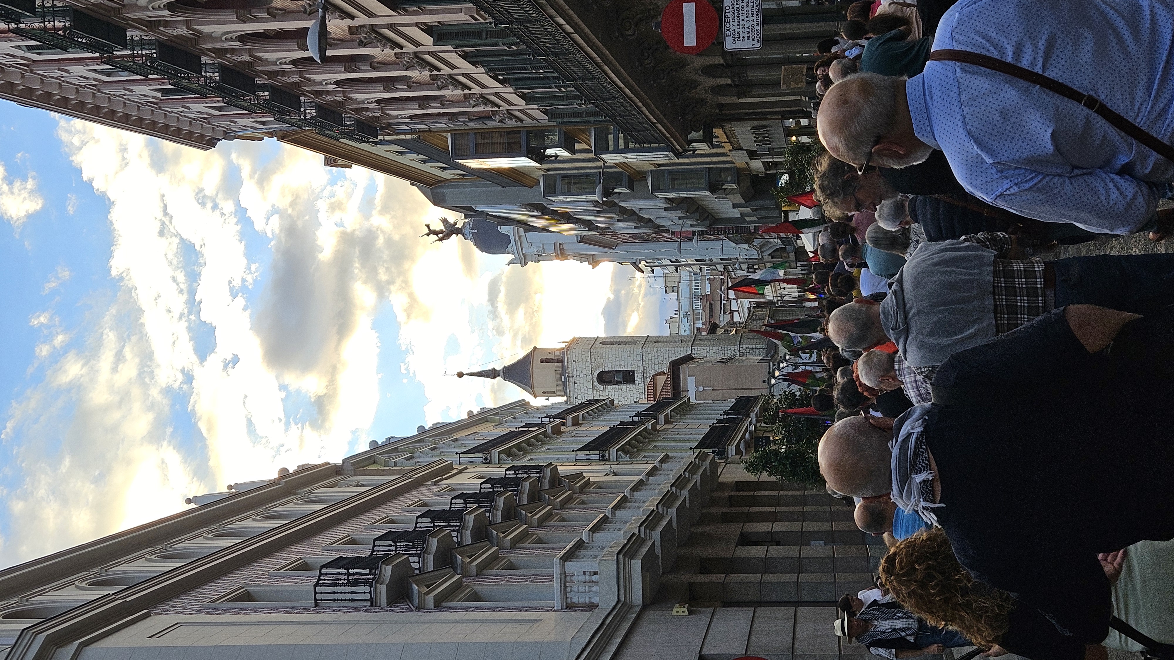 Una multitud avanza por una estrecha calle del centro histórico, entre fachadas elegantes de balcones de hierro y molduras clásicas. Al fondo, la torre de una iglesia se recorta contra un cielo de atardecer, donde la luz dorada se filtra entre nubes suaves. Entre la gente, ondean numerosas banderas palestinas que tiñen la escena. Es una manifestación en solidaridad con Palestina por las calles de Valladolid.