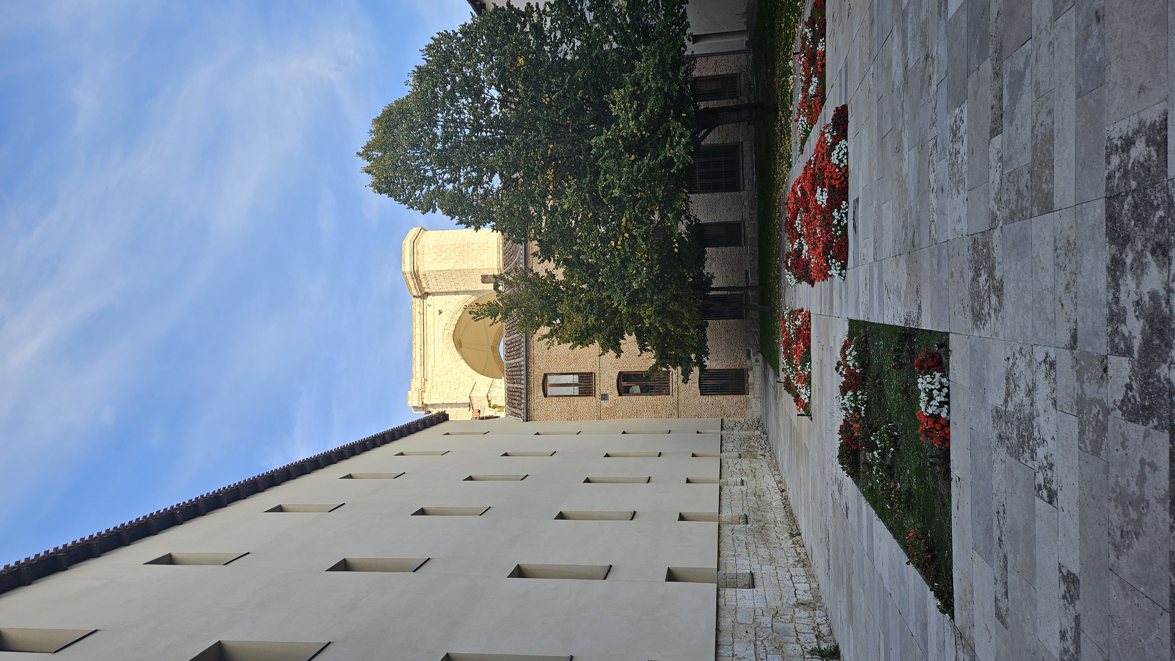 Fotografía de un patio ajardinado con parterres de flores rojas y blancas dispuestas en líneas rectas sobre un pavimento de piedra clara. A la izquierda se alza un edificio moderno de fachada beige con ventanas rectangulares, mientras que al fondo se ve una construcción más antigua de ladrillo y piedra, parcialmente oculta por dos árboles frondosos. Detrás de esta se distingue una estructura monumental de piedra clara, la iglesia de San Benito, iluminada por la luz del atardecer bajo un cielo azul con algunas nubes suaves.