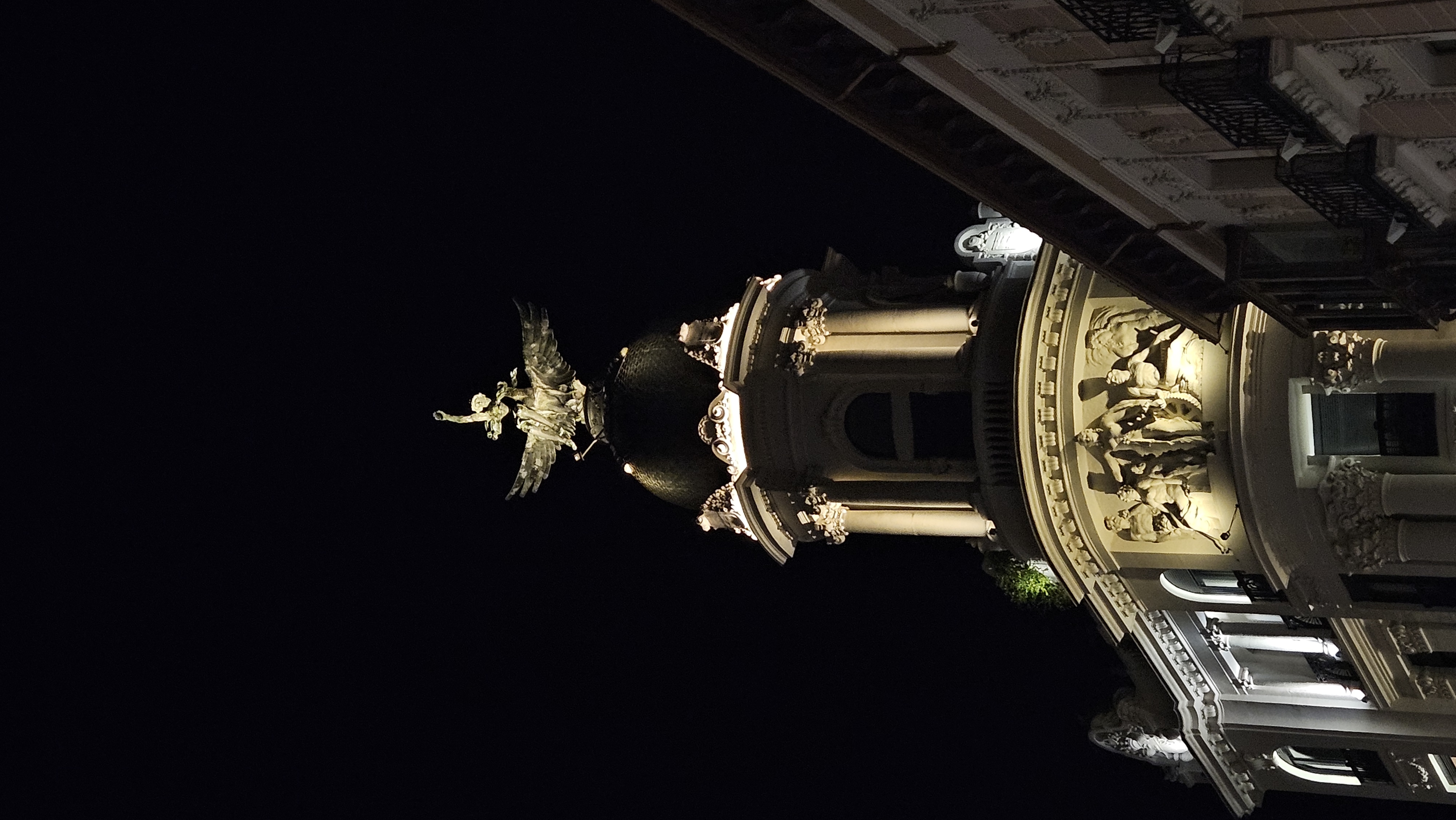 Vista nocturna de un edificio histórico en Valladolid, elegantemente iluminado. Destaca su gran cúpula oscura rematada por una escultura dorada alada que se eleva hacia el cielo. En la fachada se aprecian relieves escultóricos con figuras humanas y columnas decoradas con detalles clásicos. La iluminación resalta la textura y el relieve de la piedra, creando un contraste marcado con el cielo completamente negro y las fachadas vecinas que enmarcan la escena.