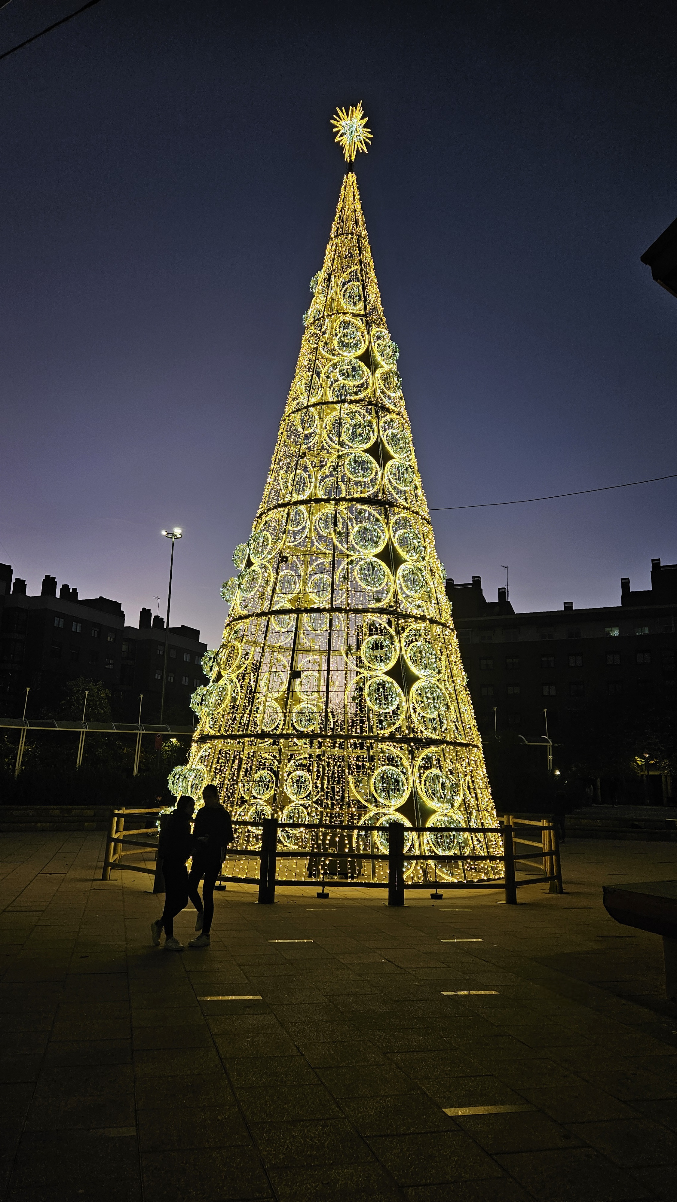 Árbol de Navidad gigante iluminado con luces doradas en una plaza al anochecer, con una pareja en silueta de pie, arrimada y de frente al árbol, y edificios residenciales oscuros al fondo bajo un cielo anocheciendo.