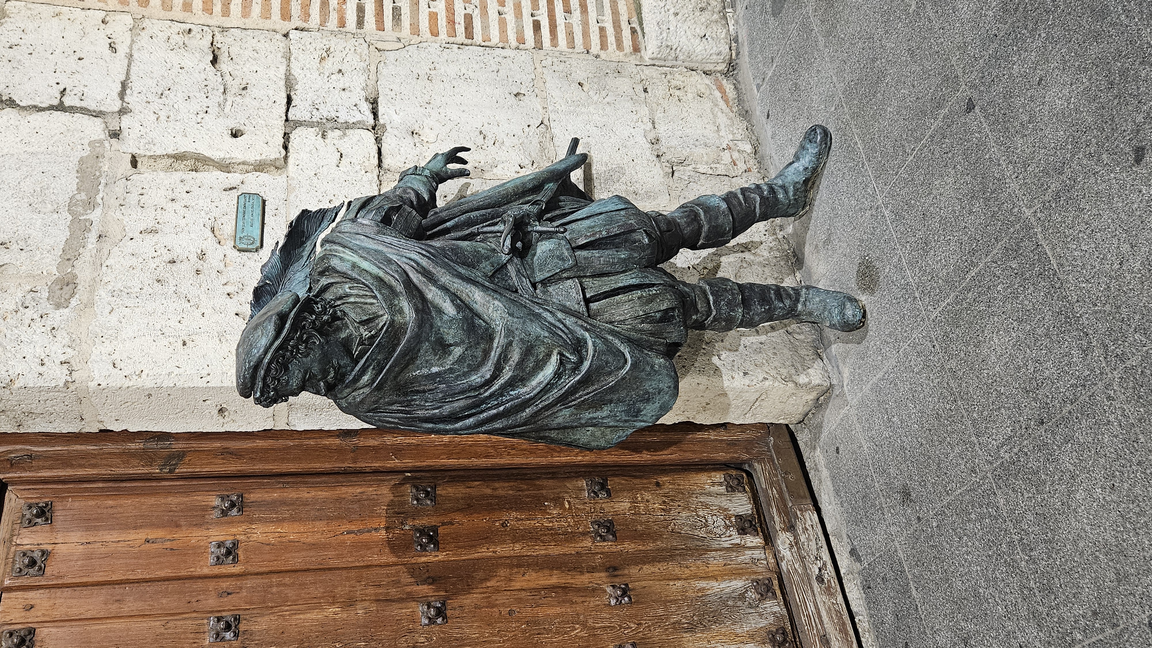 Escultura de bronce junto a la puerta de madera de la Casa de Zorrilla, en Valladolid. Representa a un caballero del Siglo de Oro, con capa, sombrero y botas altas, apoyado en la pared de piedra frente a la entrada del edificio histórico.