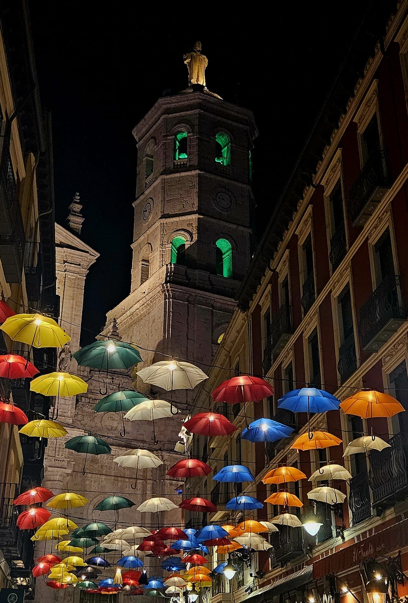 Calle nocturna en Valladolid decorada con decenas de paraguas de colores colgando en el aire e iluminados, creando un techo. Al fondo se alza la torre de la Catedral, con sus relojes y ventanales con luces verdes, coronada por una estatua iluminada de que brilla sobre el cielo oscuro. A los lados, hay edificios con balcones.