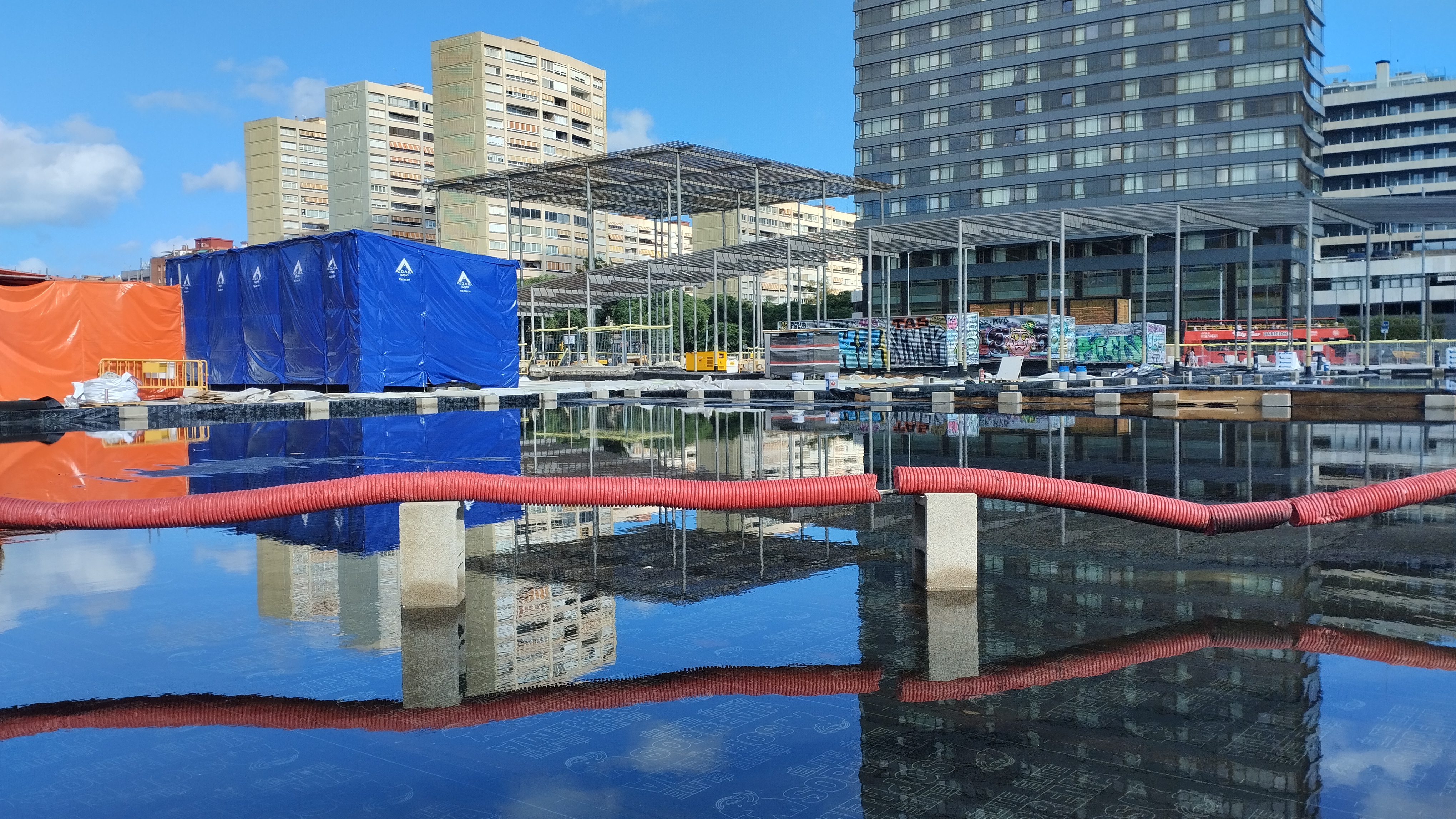 Se ve la plaza de al lado de sants en obras. concretamente se ven dos paralelepípedos uno azul y otro naranja junto con unas pérgolas aue siempre han dado mala sombra reflejadas en una lámina de agua sobre impermebeabilizante negro (con lo que casi parece un espejo). se ve también u tubo corrugado rojo atravesandolo todo sobre unos bloques de hormigón como si fuera un puente.