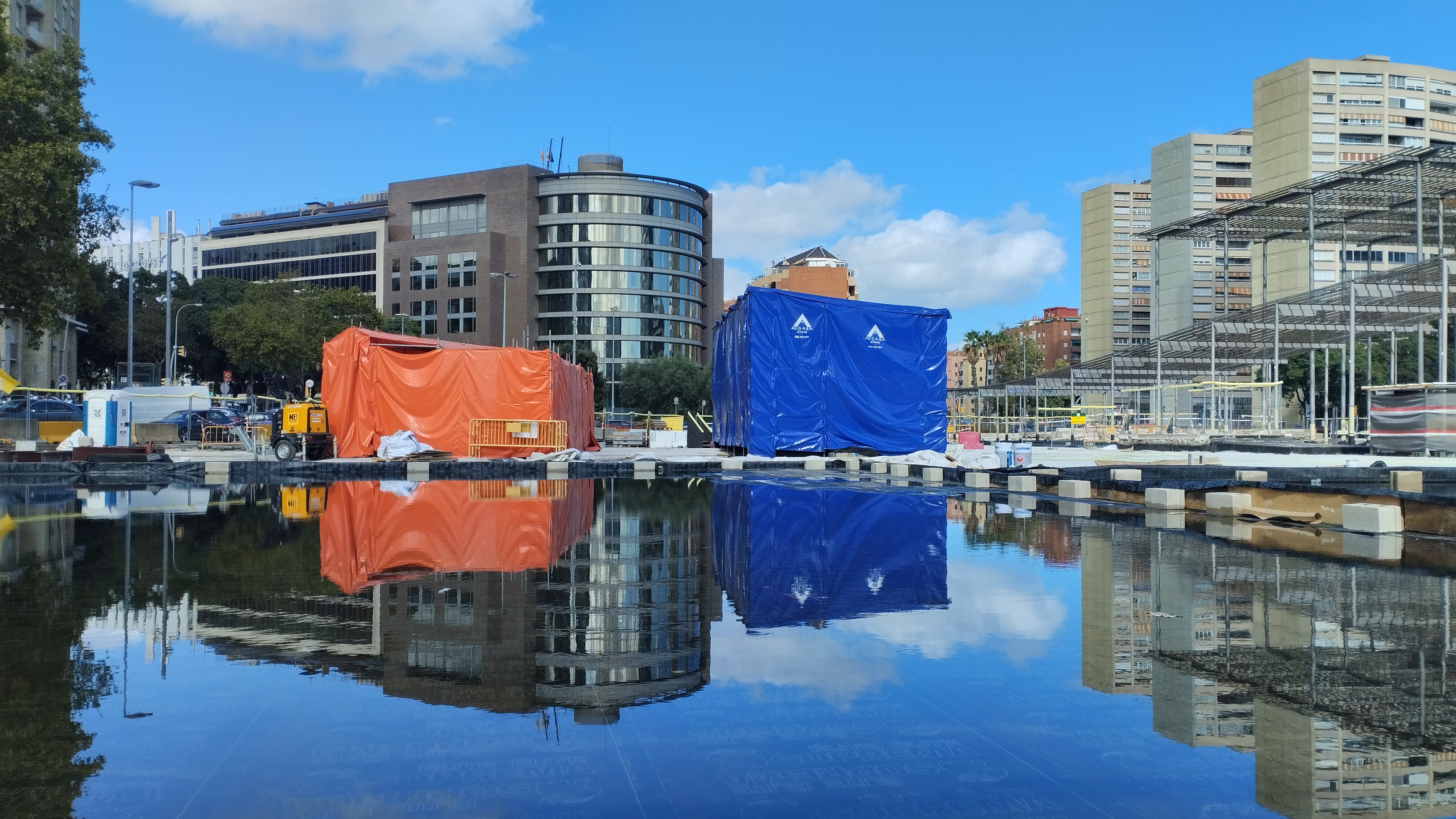 Se ve la plaza de al lado de sants en obras. concretamente se ven dos paralelepípedos uno azul y otro naranja junto con unas pérgolas aue siempre han dado mala sombra reflejadas en una lámina de agua sobre impermebeabilizante negro (con lo que casi parece un espejo).