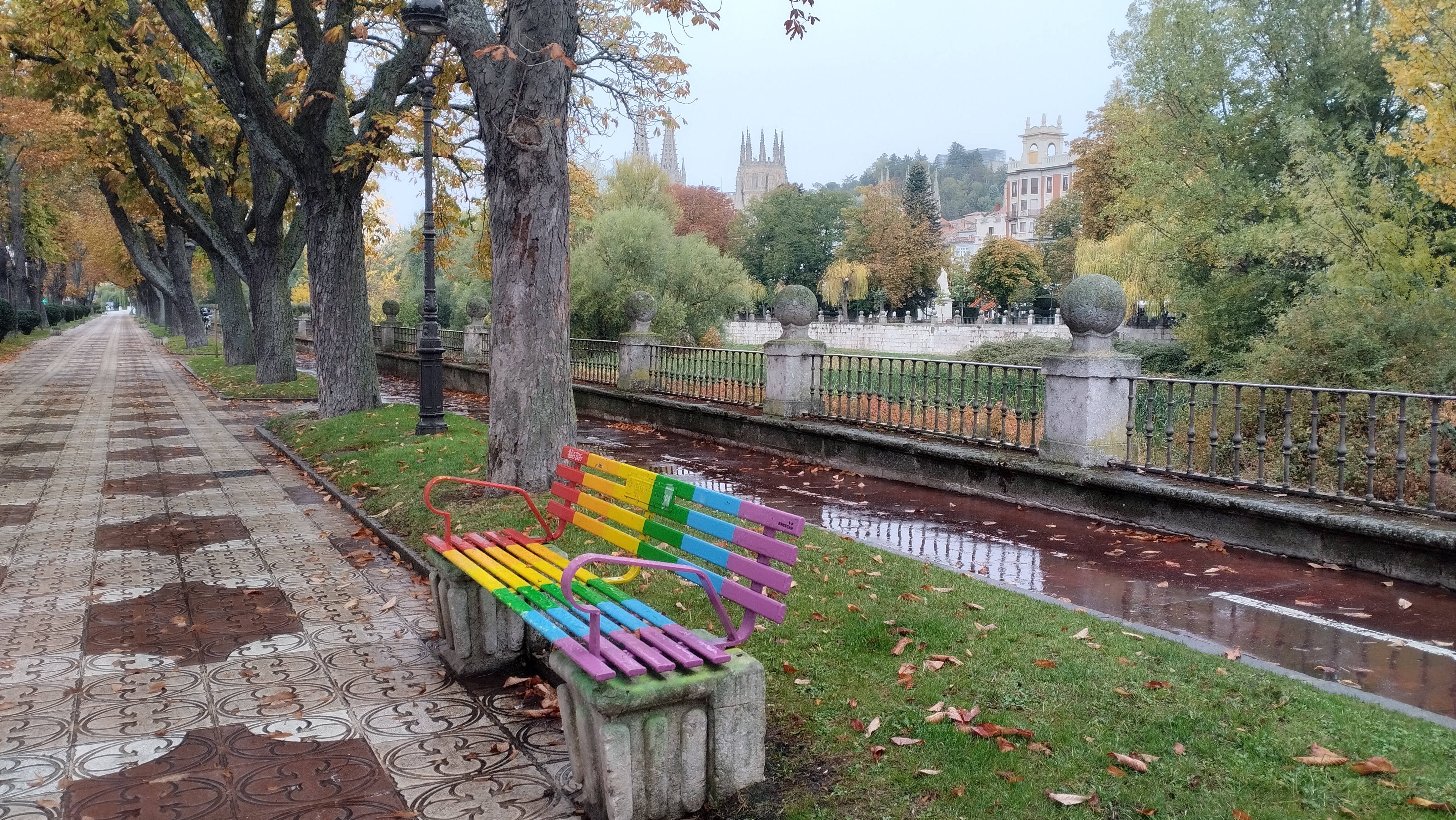 vista de la catedral desde el otro lado del río. se ve un banco pintado con colores arcoiris y los castaños con colores amarillentos