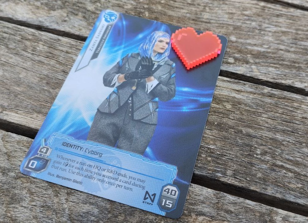 Foto de una carta de netrunner con un token con forma de corazón encima sobre un fondo que es una mesa exterior de madera.