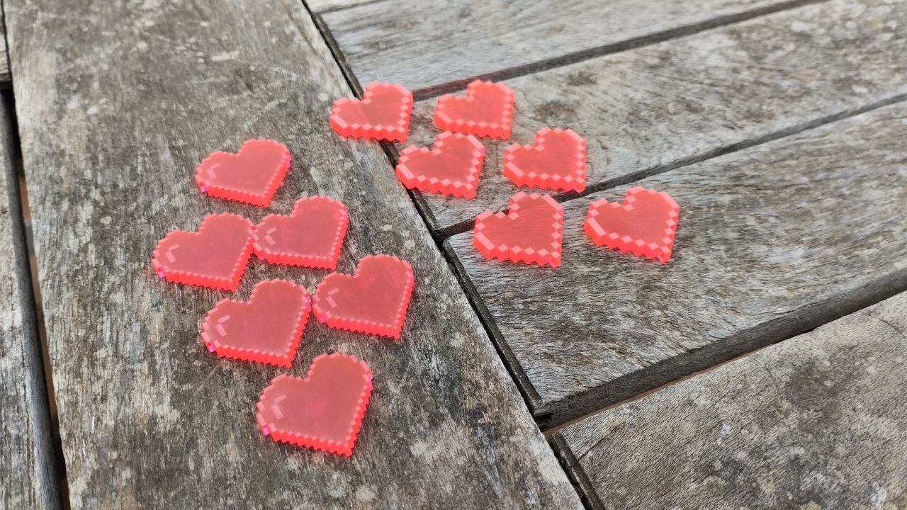 12 tokens de metacrilato con forma de corazones pixelados sobre un fondo de madera. La mitad tienen un pixel más fino que la otra mitad.